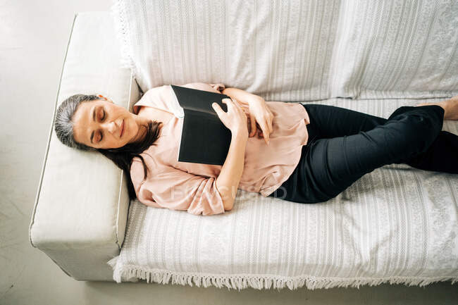 De cima de fêmea de meia-idade sonolenta deitada em sofá confortável com xadrez enquanto descansa na sala de estar em casa — Fotografia de Stock
