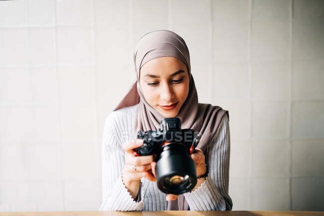 Focalizzato musulmano fotografo femminile seduto a tavola e guardando attraverso le foto sulla fotocamera professionale mentre si lavora in remoto nel caffè — Foto stock