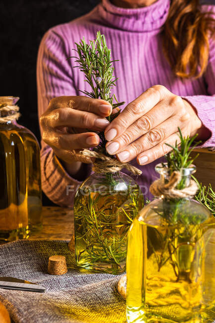 Mulher sem rosto corte em camisola roxa colocando galhos de ervas com folhas verdes em garrafa de vidro de óleo essencial perto de tesoura e corda com peito pequeno em pano na mesa de madeira — Fotografia de Stock