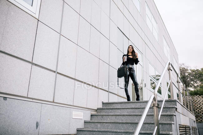 Niedriger Winkel der trendigen Unternehmerin mit Imbissgetränk im Becher steht in der Nähe moderner Gebäude auf der Stadt und blickt in die Kamera — Stockfoto
