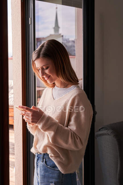 Vue latérale de la jeune femme calme dans la messagerie pull élégant sur smartphone tout en se tenant près de la fenêtre à la maison — Photo de stock