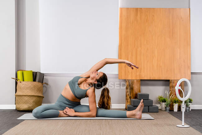 Full body of flexible calm young lady in sportswear stretching spine in Revolved Head to Knee pose while practicing yoga in studio near smartphone on tripod and laptop — Stock Photo