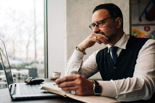 Pensive mature ethnic male executive touching mouth at table with netbook and diary in cafeteria — Stock Photo