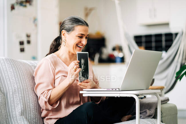 Side view of smiling middle aged female sitting on couch and showing picture of baby while having video chat on netbook on blurred background — Stock Photo