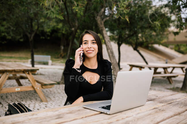 Усміхнена жінка-підприємець сидить за столом з ноутбуком в парку і виступає на смартфоні, працюючи віддалено і озираючись — стокове фото