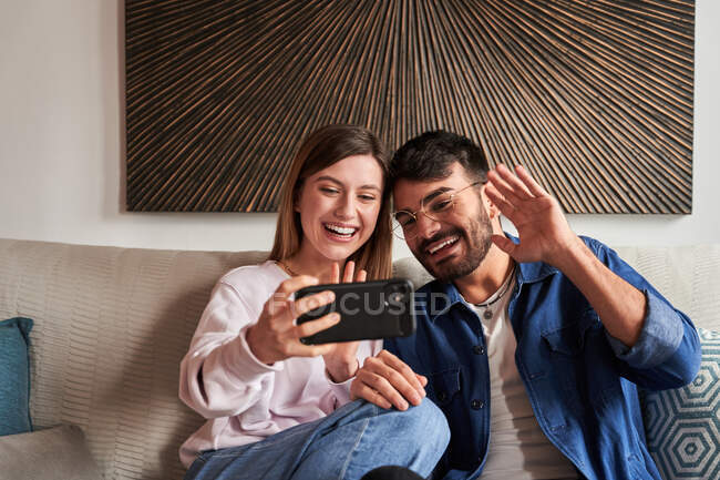 Romântico jovem casal diversificado em roupas casuais relaxando no sofá e assistindo fotos no smartphone durante o fim de semana em casa — Fotografia de Stock