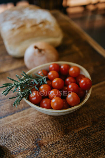Високий кут миски зі свіжими вишневими помідорами біля стовбурів розмарину і цілої цибулі на дерев'яному столі — стокове фото