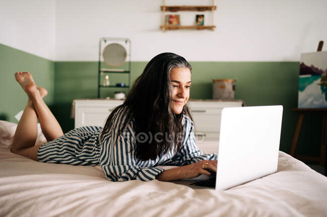 Боковой вид позитивной зрелой самозанятой женщины с длинными темными волосами в повседневной одежде, лежащей на кровати и сидящей на ноутбуке во время онлайн-работы дома — стоковое фото