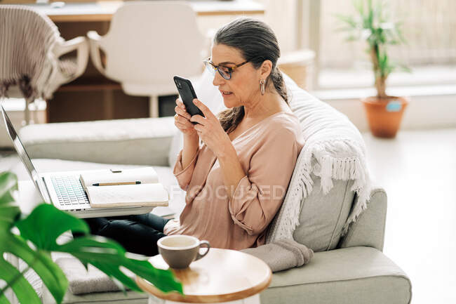 Vista lateral da navegação feminina concentrada de meia-idade no smartphone enquanto trabalhava no laptop na sala de luz em casa — Fotografia de Stock