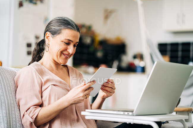 Fröhliche Frau mittleren Alters sitzt auf einer Couch in der Nähe moderner Netbooks, während sie sich ein Foto in einer modernen Wohnung mit Küche auf verschwommenem Hintergrund ansieht — Stockfoto