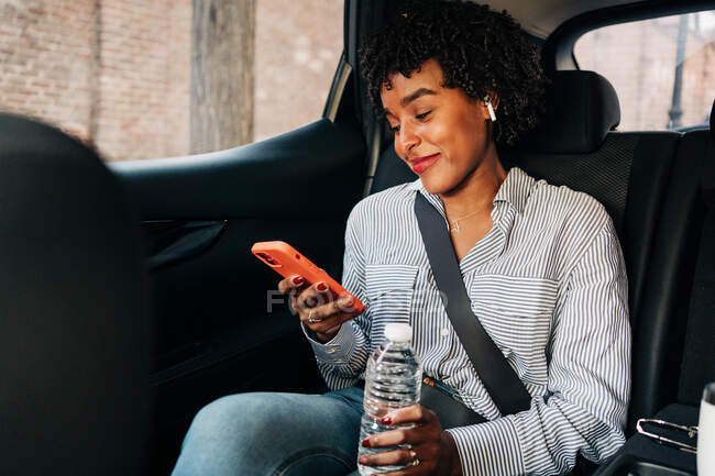 Cheerful young African American female in striped shirt browsing mobile phone while riding in car having bottle of water — Stock Photo