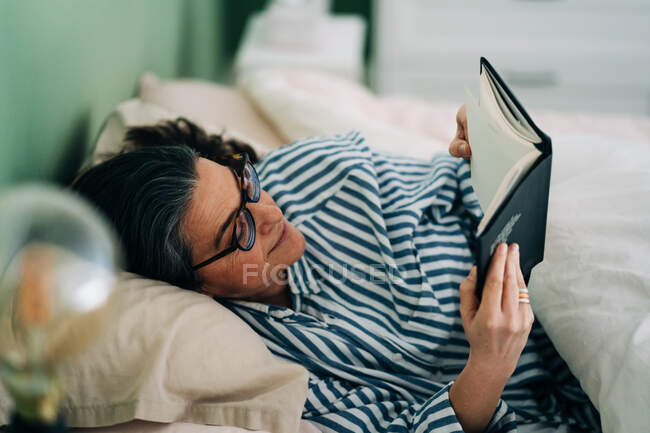 Relajada mujer hispana madura en ropa casual y anteojos acostada en la cama y leyendo interesante libro antes de dormir - foto de stock