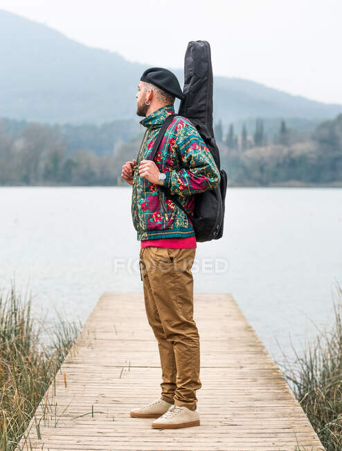 Vista lateral de macho pensativo em roupas elegantes de pé com guitarra em caso de no cais de madeira perto do rio calmo contra a montanha em dia nublado — Fotografia de Stock