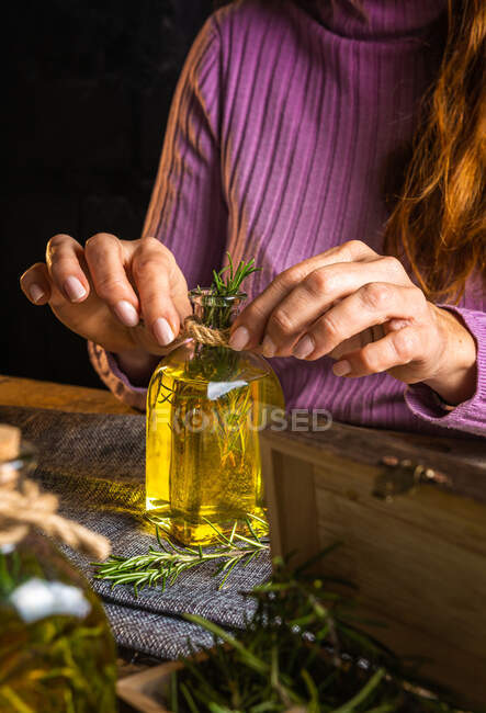 Crop señora anónima en suéter púrpura que muestra botellas de vidrio de aceite esencial con ramitas de hierbas con hojas verdes cerca de tela en la mesa - foto de stock