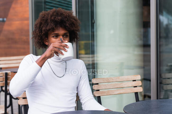 Relaxed young black guy with Afro hairstyle in stylish clothes drinking coffee and looking away while sitting on cafe terrace in city — Stock Photo