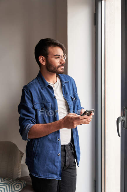 Visão lateral de pensivo jovem barbudo macho étnico em roupas casuais e óculos de pé perto da janela e mensagens no telefone celular em casa — Fotografia de Stock