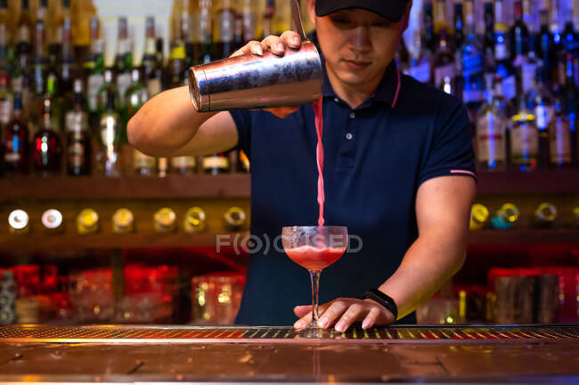 Молодой азиатский бармен работает в баре с шейкером и наливает коктейль в стакан — стоковое фото