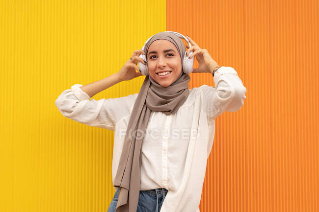 Mulher étnica alegre em lenço de cabeça e fones de ouvido sem fio ouvindo música à luz do dia — Fotografia de Stock