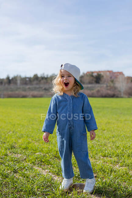Corpo cheio de menina feliz em roupas elegantes e boné olhando para longe, enquanto em pé na grama no dia ensolarado de verão no campo — Fotografia de Stock
