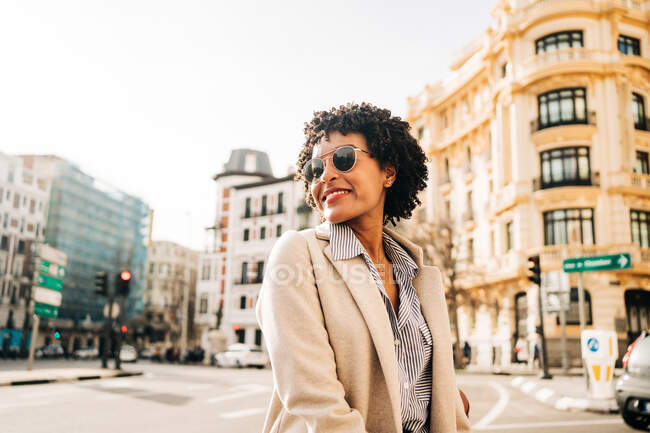Молода афроамериканська жінка в модному одязі посміхається і дивиться на міську вулицю в сонячному світлі — стокове фото