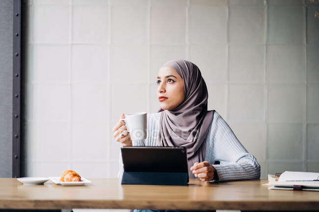 Mujer musulmana freelancer pensativa en pañuelo para la cabeza sentada en la mesa con una taza de bebida y una tableta mientras piensa en el proyecto y mira hacia otro lado - foto de stock
