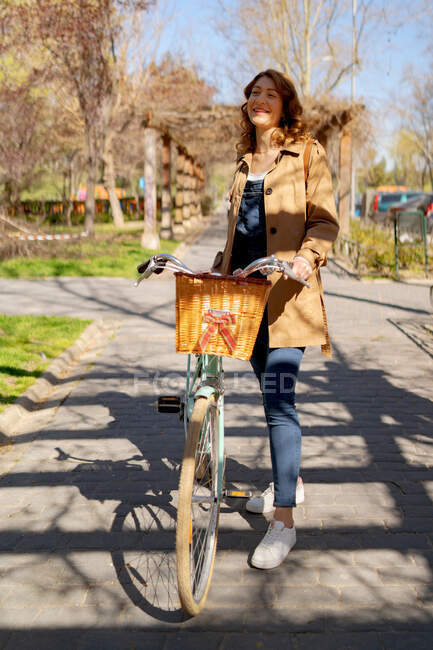 Cuerpo completo de alegre joven hembra sonriendo y mirando hacia otro lado cerca de la vieja bicicleta con cesta de mimbre de madera - foto de stock