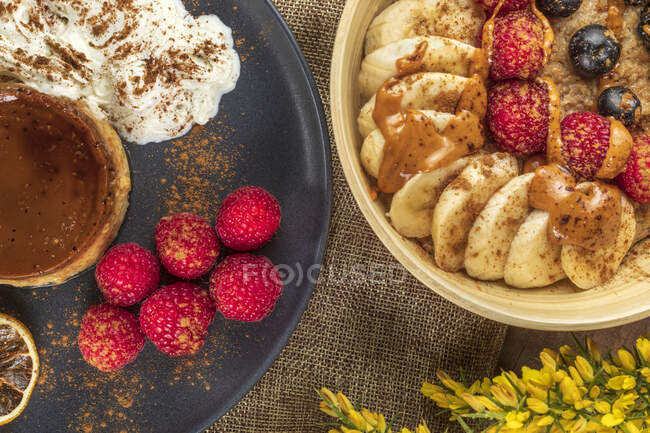 From above of tasty flan with whipped cream and fresh raspberries near bowl full of banana slices and berries with caramel sauce — Stock Photo