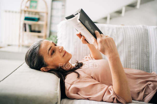 Vista lateral do livro de leitura feminina enquanto deitado no sofá confortável na sala de estar com planta verde em casa — Fotografia de Stock