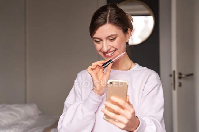 Felice giovane blogger donna in abiti casual sorridente e dimostrando lucidalabbra durante le riprese video su smartphone per vlog — Foto stock