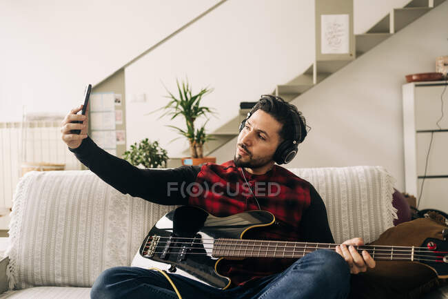 Unshaven male artist in headset with electric guitar taking self portrait on cellphone while sitting on couch in house — Stock Photo