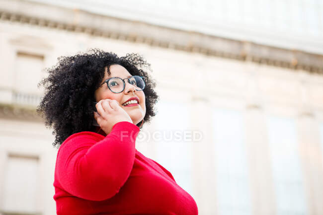 Знизу вмісту надмірної ваги жінка в червоному одязі та окулярах розмовляє на мобільному телефоні, дивлячись вгору по місту — стокове фото