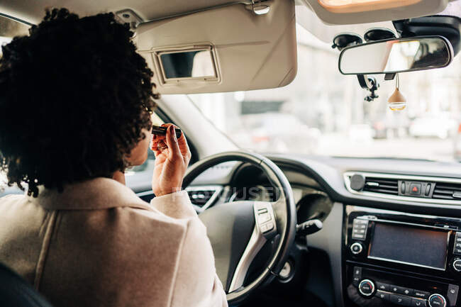 Rückansicht einer nicht wiederzuerkennenden afroamerikanischen Fahrerin, die im Auto sitzt und Lippenstift aufträgt, während sie in den Spiegel schaut — Stockfoto