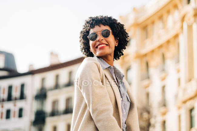 Joven mujer afroamericana feliz en traje de moda sonriendo y mirando hacia otro lado en la calle urbana bajo el sol - foto de stock
