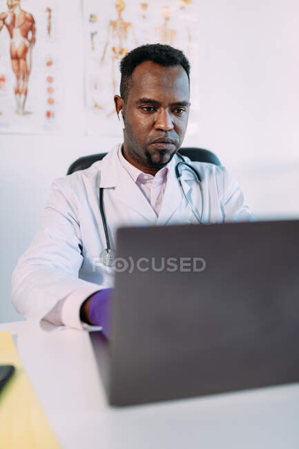 Médico masculino afro-americano concentrado em robe médico e fones de ouvido TWS trabalhando em laptop enquanto sentado à mesa na clínica moderna — Fotografia de Stock
