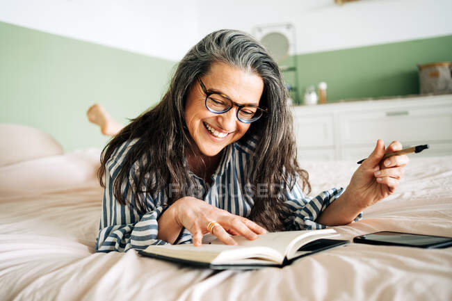 Middle aged cheerful female in striped shirt smiling and reading notebook on bed with smartphone — Stock Photo