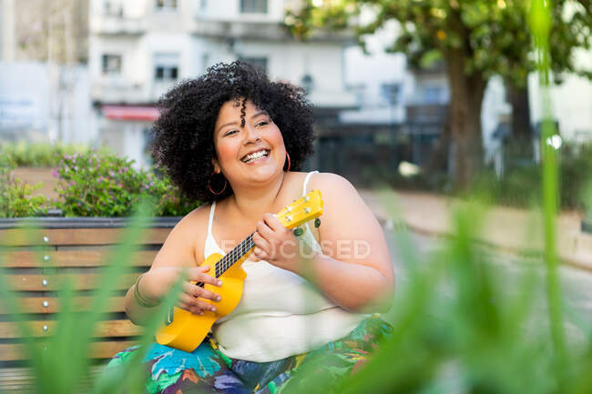 Fröhliche Künstlerin in Zierkleidern spielt Musikinstrument auf Stadtbank — Stockfoto