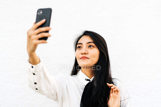 Mulher asiática com cabelos longos e escuros tomando auto retrato no celular em pé no fundo branco — Fotografia de Stock