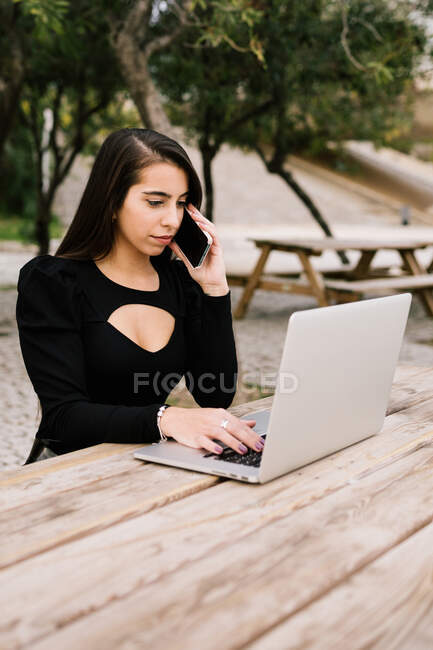 Жінка-підприємець сидить за столом з ноутбуком в парку і виступає на смартфоні під час роботи віддалено — стокове фото