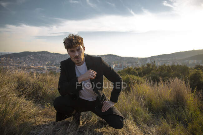Adult bearded male in coat squatting on faded grass and looking at camera against mounts under cloudy sky in twilight — Stock Photo
