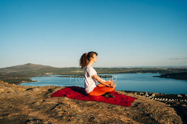 Vue latérale de la jeune femme aux longs cheveux bouclés en vêtements de sport méditant avec les yeux fermés dans la pose Lotus assis sur une pente rocheuse au-dessus de la mer sous un ciel bleu sans nuages — Photo de stock