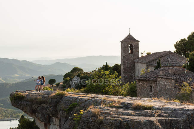 Удаленный обзор женщин-туристок, стоящих на краю скалы и делающих автопортрет на фоне древнего замка, расположенного в высокогорье — стоковое фото