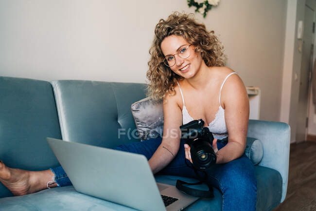 Весела молода жінка-блогер з кучерявим світлим волоссям в повсякденному вбранні сидить на дивані і посміхається при передачі фотографій з професійної камери на ноутбук — стокове фото
