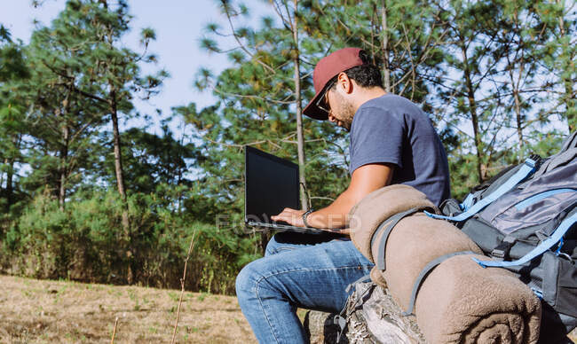 De baixo nómada sentado em pedra perto de pertences e usando netbook enquanto trabalhava remotamente — Fotografia de Stock