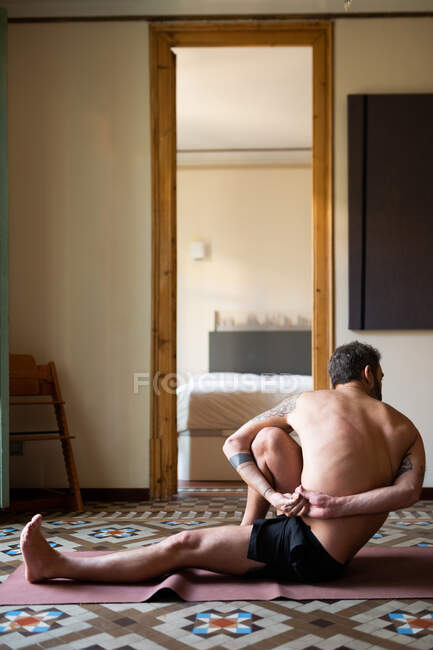 Vista lateral de macho con torso desnudo practicando yoga sobre estera en Marichyasana en casa - foto de stock