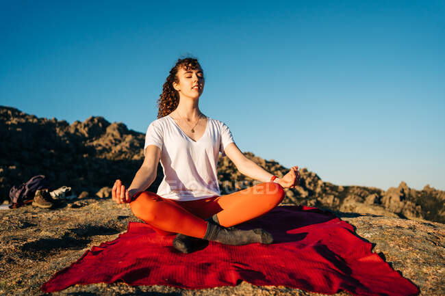 Young lady with long curly hair in activewear meditating with closed eyes in Lotus pose sitting on rocky hill slope under cloudless blue sky — Stock Photo