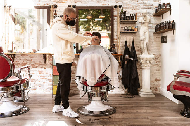 Peluquería masculina en máscara textil recortando pelo de cliente anónimo en sillón contra espejo en barbería - foto de stock