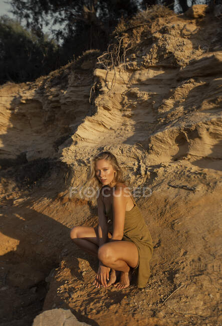 Full body of barefoot sensual blond female sitting on haunches near sandy slope with grass plants — Stock Photo