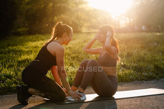 Side view of slim female athlete doing abdominal crunches with help of sportswoman during training at sunset in park — Stock Photo