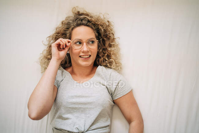 Vista superior de la joven confiada con el pelo rubio rizado en ropa casual ajustando las gafas y sonriendo mientras está acostado en la cama en casa - foto de stock