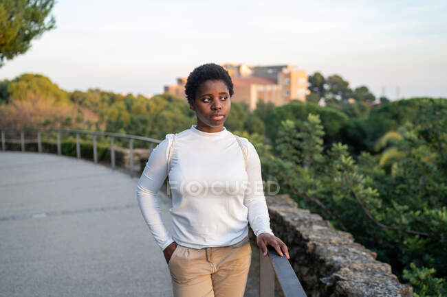 Молода афроамериканська жінка в повсякденному одязі стоїть біля паркану в пишному міському парку в літній день і озирається геть — стокове фото